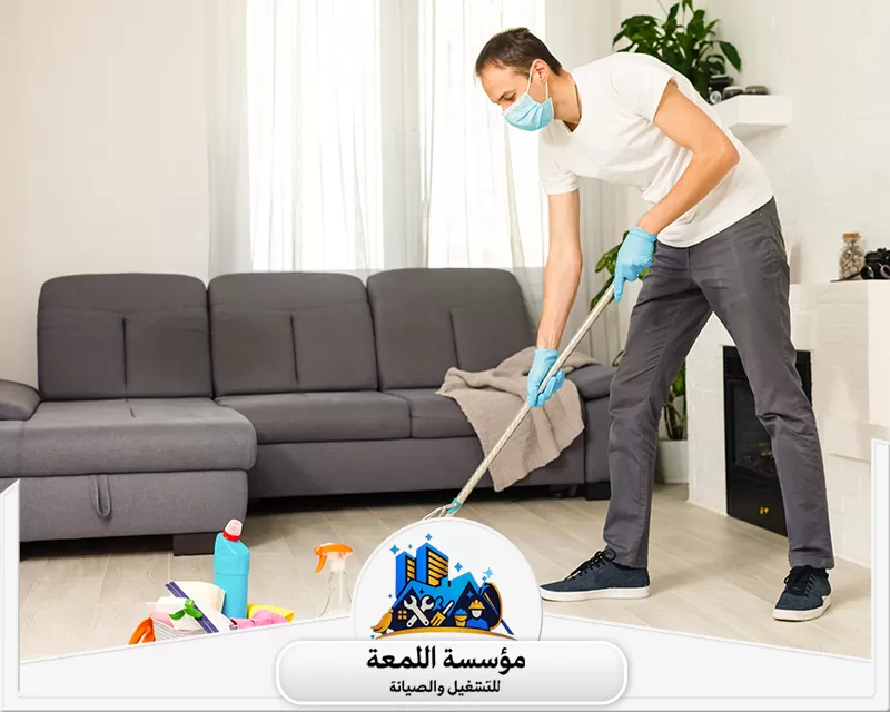 شركة تنظيف بالمجمعة 0565283428 شركة اللمعة تنظيف مسح أرضيات داخلية بممسحة. عامل يرتدي كمامة وقفازات، مع معدات تنظيف في الخلفية لضمان النظافة.